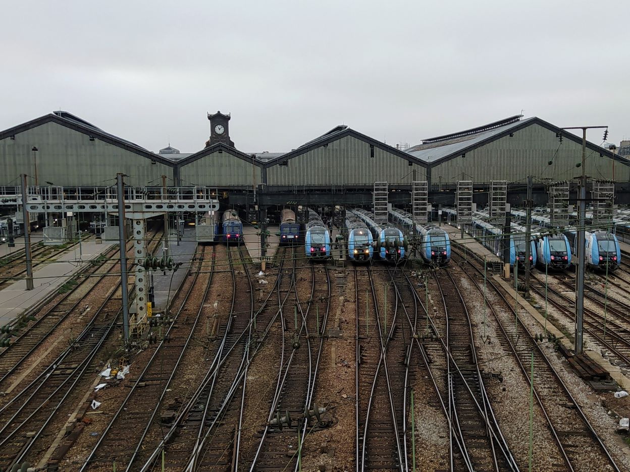 Saint-Lazare station from behind. We can see 15 tracks, most of which have trains in them in Île-de-France Mobilités livery. Behind them is the station roof, above which stands a small clock tower